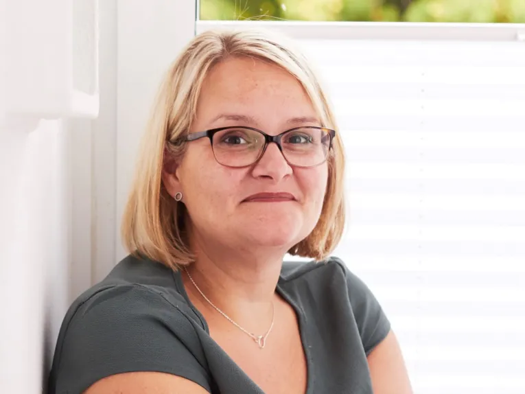 Eine Person in einem grauen Oberteil und einer Brille l&auml;chelt und schaut in die Kamera.