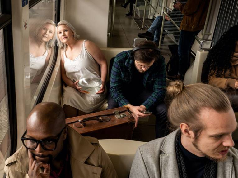 Schauspielerin im Nachthemd und mit einem Goldfischglas in einer hannoverschen Stadtbahn mit anderen Fahrgästen.