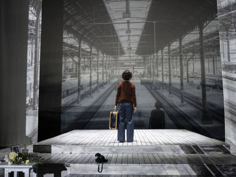 Eine Schauspielerin steht mit einem Koffer in der Hand auf einer Bühne, auf deren Wände Bahnsteige eines Bahnhofs projiziert sind.