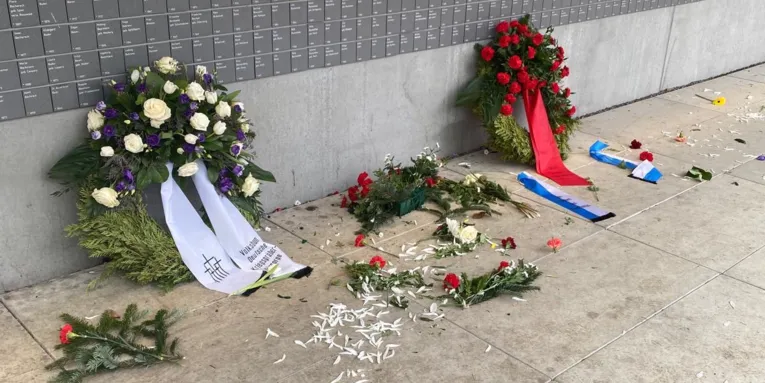 Teile zerstörter Kränze liegen an der Wand der Namen oder im Beet davor.