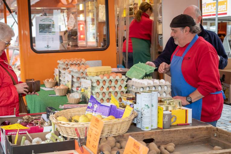 Ein Verkäufer und eine Kundin an einem Stand an dem unter anderem Eier und Gemüse verkauft werden.
