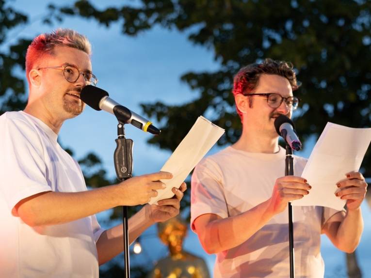 Zu sehen sind zwei Poetry Slamer auf einer Openair Bühne, während sie aus ihren texten vorlesen.
