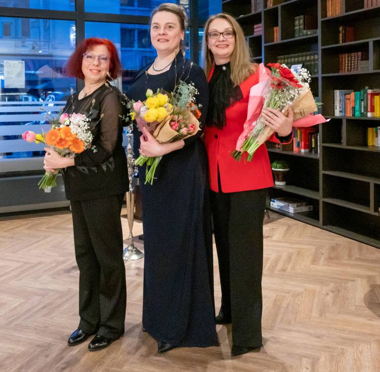 Drei Frauen in Abendgarderobe stehen vor einem Regal und halten Blumen in den Armen.