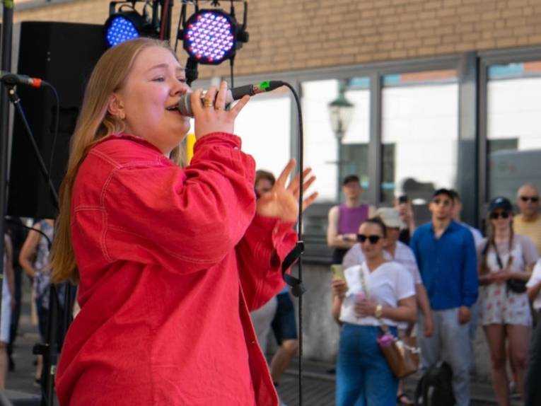 Frau in roter Kleidung, die vor mehreren Menschen ein Mikrofon zum Mund hinhält und singt