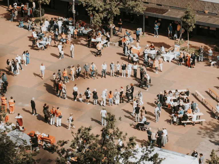 Blick auf einen Platz, auf dem Menschen stehen und an Tischen Sitzen. In der Mitte bilden Menschen einen Kreis und halten sich teilweisen an den Händen.