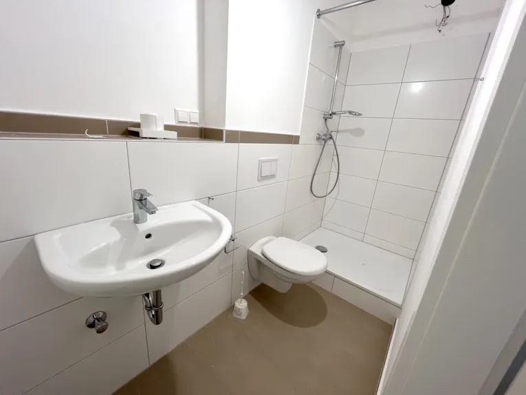 Blick in einen kleinen wei&szlig; gefliesten Raum mit einem Waschbecken, einer Toilette und einer Dusche