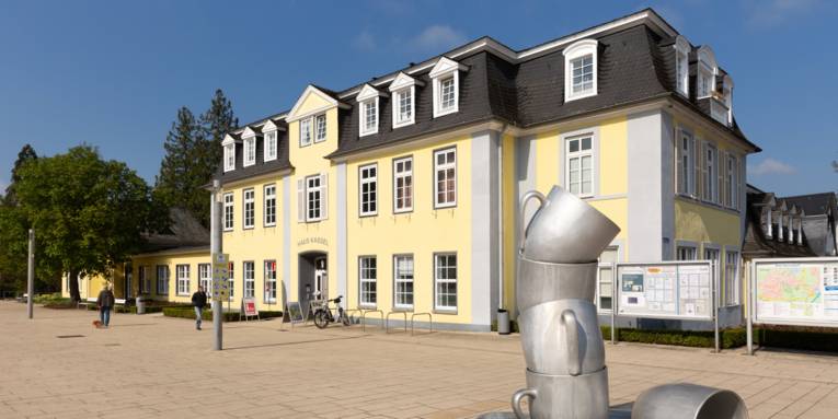 Gelbe Villa mit grüner Eingangstür und gepflastertem Fußgängerbereich. Davor ist ein Kunstwerk, das aussieht, wie ein überdimensionaler Stapel aus Geschirr.