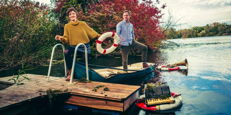 Ein Mann und eine Frau halten einen Rettungsring, während sie mit einem Boot an einem Steg anlegen. Im Wasser treiben Instrumente in weiteren Rettungsringen.