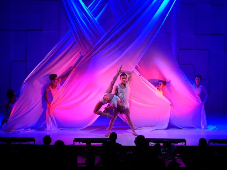 Tänzer in weißen Outfits performen vor kunstvoll drapierten Stoffbahnen, beleuchtet in blauem und pinkem Licht.