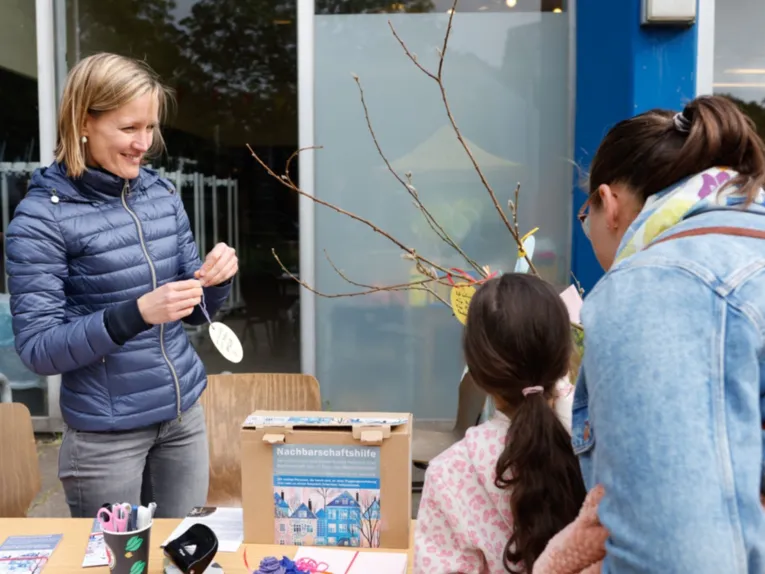 Ein M&auml;dchen und eine Frau stehen vor einem Tisch mit einem Karton, auf dem Nachbarschaftshilfe steht. Hinter dem Tisch steht eine Frau und l&auml;chelt.