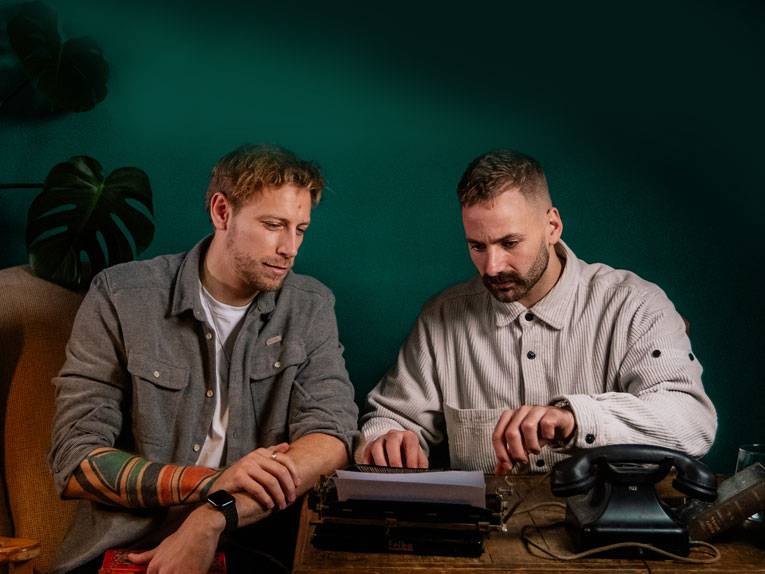 Zwei Männer sitzen an einem Schreibtisch, auf dem eine Schreibmaschine und ein Telefon stehen