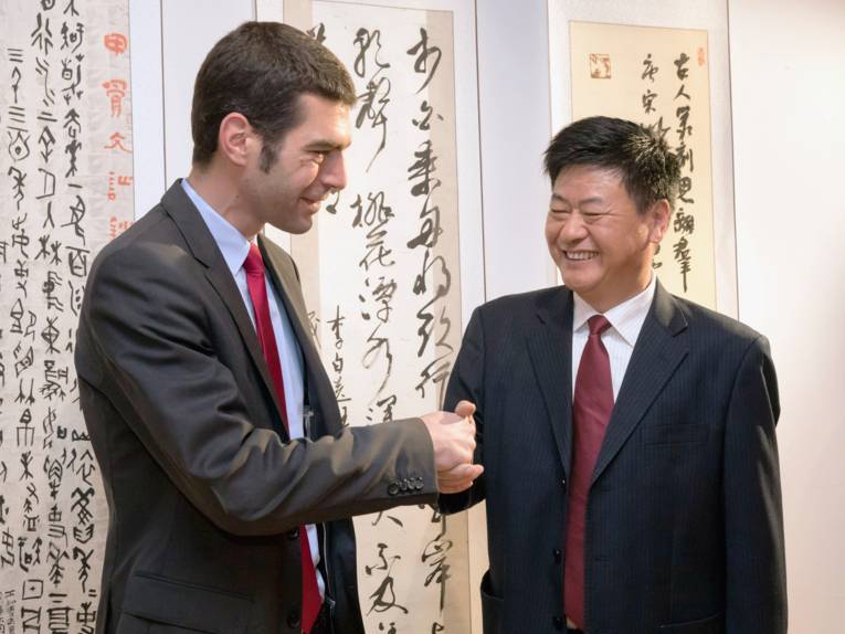 Zwei Männer reichen sich die Hände, hinter ihnen hängen weiße Papierbahnen mit chinesichen Schriftzeichen in roter und weißer Farbe.