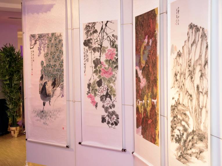 Vier Beispiele chinesischer Malerei hängen an einer Ausstellungswand, die Bilder zeigen einen Pfau, Blüten und zwei Berglandschaften.