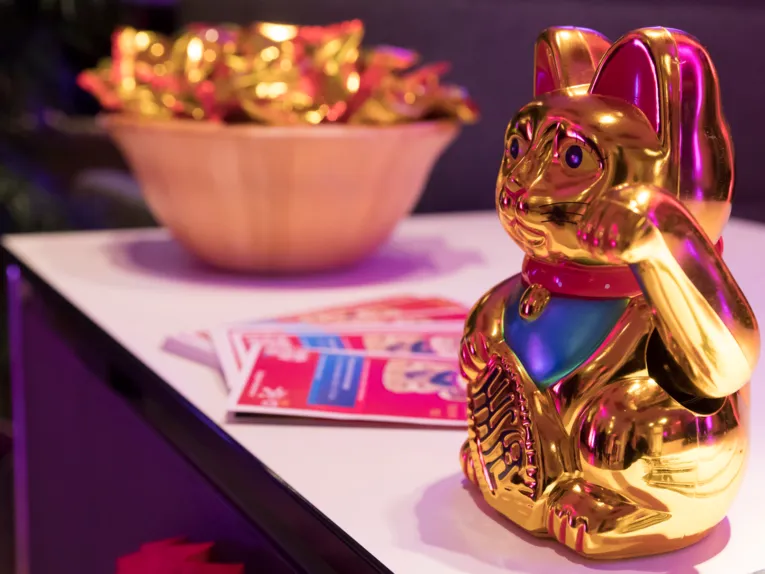 Figur einer goldenen Katze mit rotem Halsband steht auf einem Tisch, außerdem sind Flyer und eine Schale mit Glückskeksen auf dem Tisch.