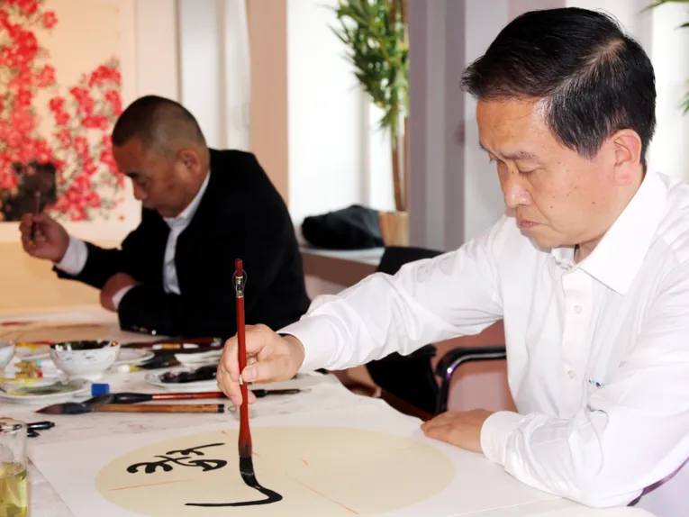 Ein Mann trägt schwarze Farbe mit einem Pinsel auf Papier auf, es entstehen chinesische Schriftzeichen.