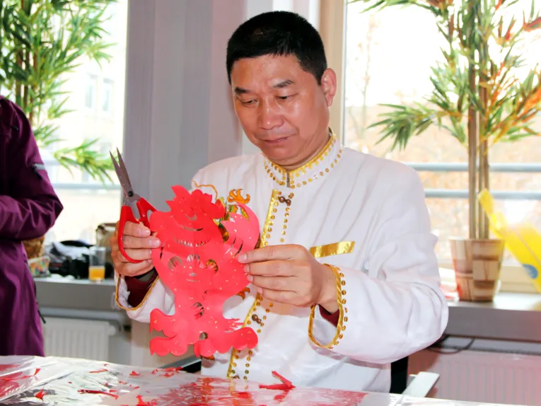 Ein Mann bearbeitet Papier mit einer Schere, es entsteht ein roter Hahn.