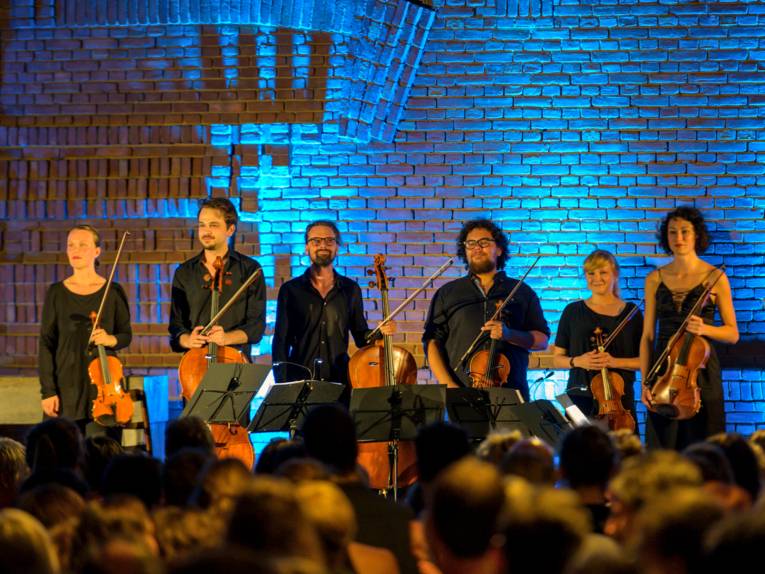 Musikerinnen und Musiker stehen mit ihren Instrumenten vor einem beleuchteten Hintergrund.