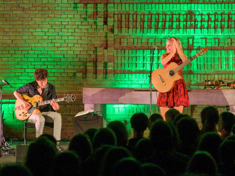 Ein Musiker und eine Musikerin stehen mit Gitarran auf einer grün beleuchteten Bühne.