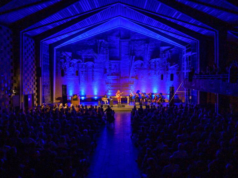 Blick von den hinteren Reihen einer Kirche hin zu einer blau beleuchteten Bühne.