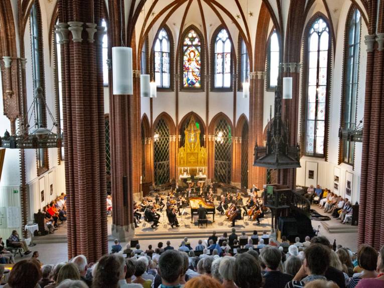 Blick aus den Reihen der Zuschauerinnen und Zuschauer auf ein Orchester, das in einer Kirche auftritt.