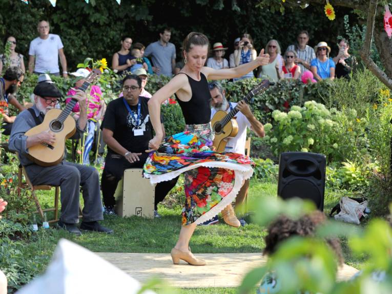 Eine Frau tanzt auf einer improvisierten Tanzfläche in einem Schrebergarten, dazu spielen hinter ihr drei Musiker, dahinter betrachtet Publikum die Szene.