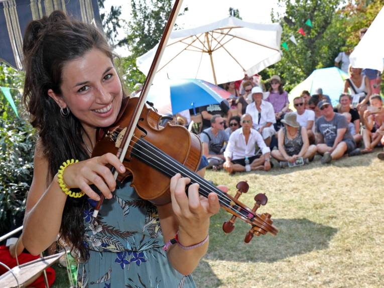 Eine Violonistin spielt auf ihrem Instrument und schaut in die Kamera, dahinter ist Publikum auf einer Picknickwiese.