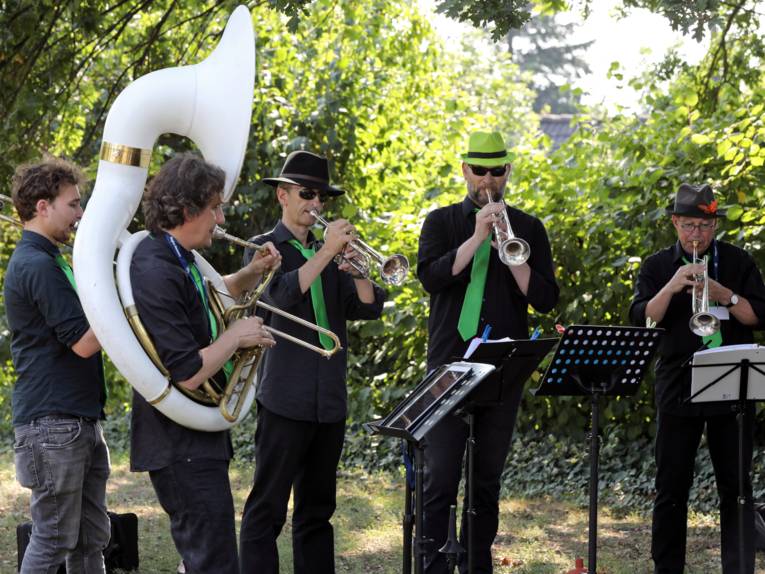 Fünf Musiker spielen unter freiem Himmel und in einer grünen Kulisse auf ihren Instrumenten.