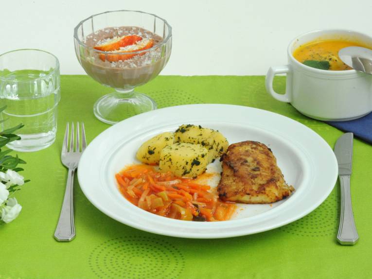Ein Teller mit Kartoffeln, Gemüse und einem Stück Fleisch, am Rande eine Tasse Suppe, ein Glas Wasser und ein Dessert