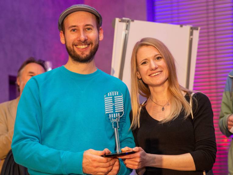 Ein Mann und eine Frau, beide halten einen gläsernen Pokal in Form eines Mikrofons in den Händen