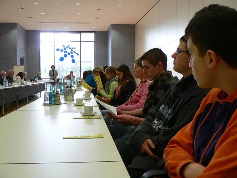 Schülerinnen und Schüler am einm langen Konferenztisch an dessen Ende eine Frau steht