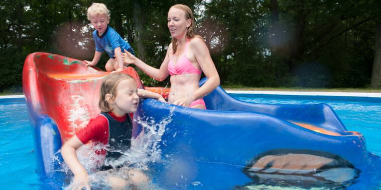 Frau mit zwei Kindern an einem Spielgerät, dass in einem Nichtschwimmerbereich eines Freibades steht