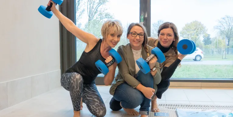 Drei Damen, teils in sportlicher Kleidung, sitzen am Rand eines Schwimmbeckens und halten blaue Hanteln in den Händen.