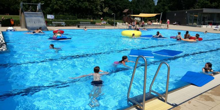 Kinder bei Sonnenschein mit großen blauen Matten und großem gelben Reifen im Außenschwimmbecken, im Hintergrund eine breite Edelstahl Rutsch