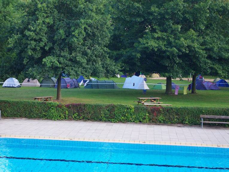 Zeltlager auf der einer mit Bäumen bestandenen Wiese, im Vordergrund Teile eines Schwimmbecken