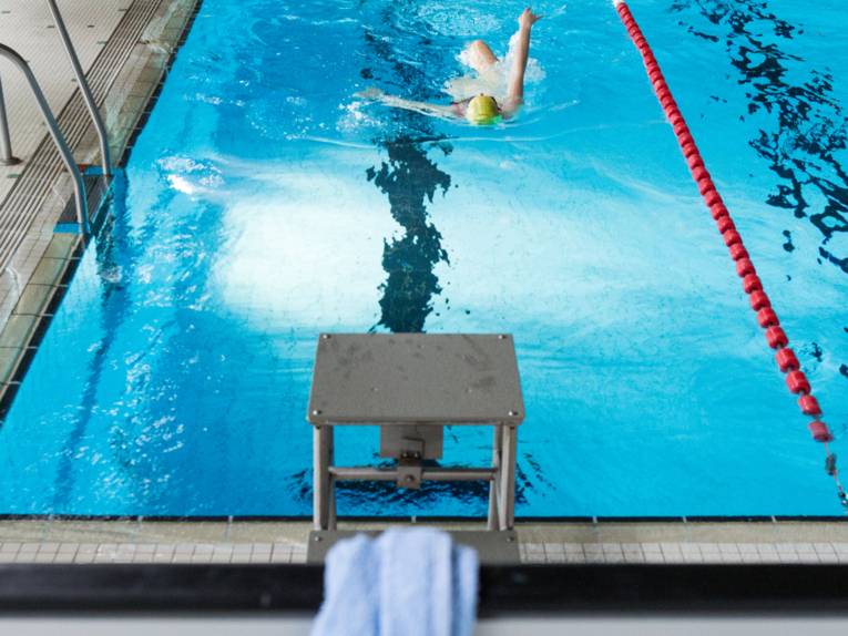 Person, die im Hallenbad auf einer Schwimmbahn Rückenschwimmen macht. Im Vordergrund ein Startblock mit Handtuch.