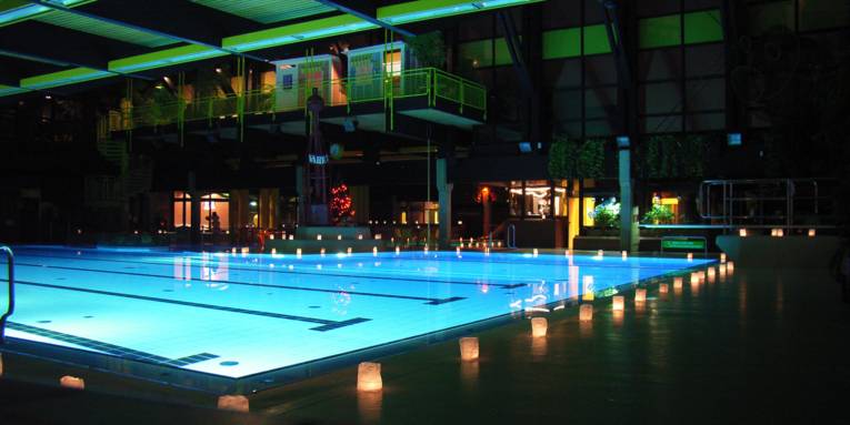 Beleuchtetes Schwimmbecken eines Hallenbades am Abend, am Rand stehen weisse, leuchtende Tüten
