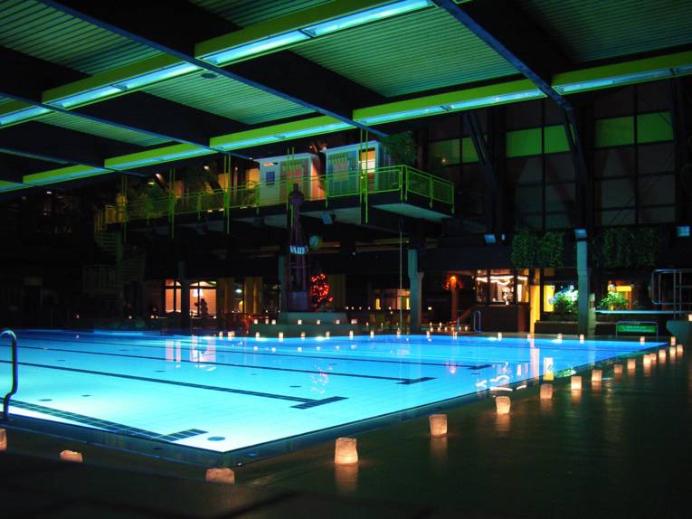 Beleuchtetes Schwimmbecken eines Hallenbades am Abend, am Rand stehen weisse, leuchtende Tüten