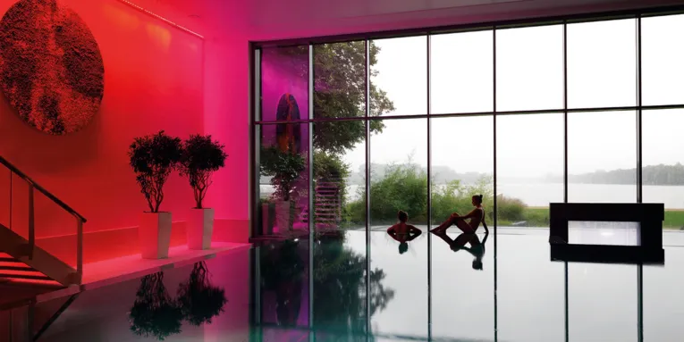 Blick über die Wasserfläche eines Schwimmbeckens, am hinteren Beckenrand sind zwei junge Frauen, dahinter ist eine große Fensterfläche. Durch die Fenster ist der Maschsee zu sehen.