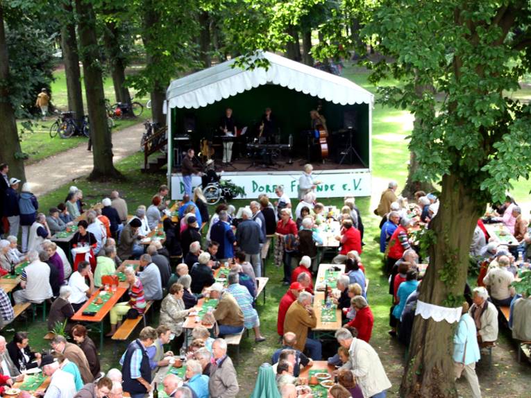 Eine kleine Bühne ist unter Bäumen aufgebaut, darauf spielen Musiker Jazz. Davor sitzen viele Besucherinnen und Besucher auf Festzeltgarnituren.
