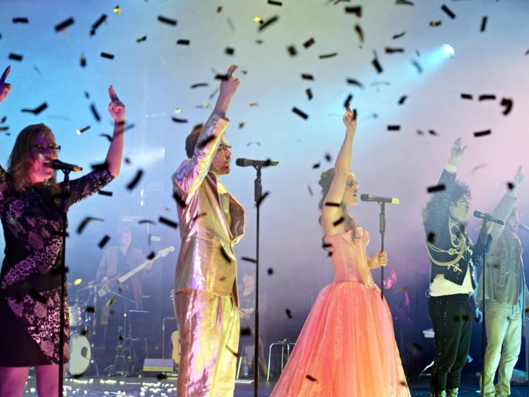 Fünf Schauspieler stehen auf einer Bühne, von oben fällt Flitter herunter.