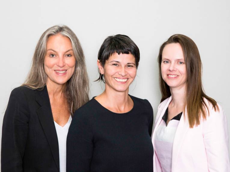 Drei Damen lächeln vor einem grauen Hintergrund in die Kamera.