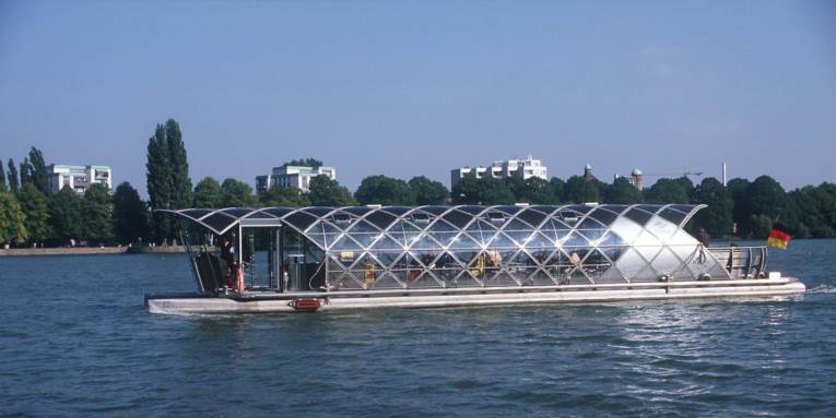 Boot mit Glasdach auf einem Gewässer, im Hintergrund Bäume und mehrstöckige Häuser