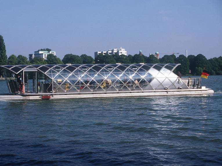 Boot mit Glasdach auf einem Gewässer, im Hintergrund Bäume und mehrstöckige Häuser