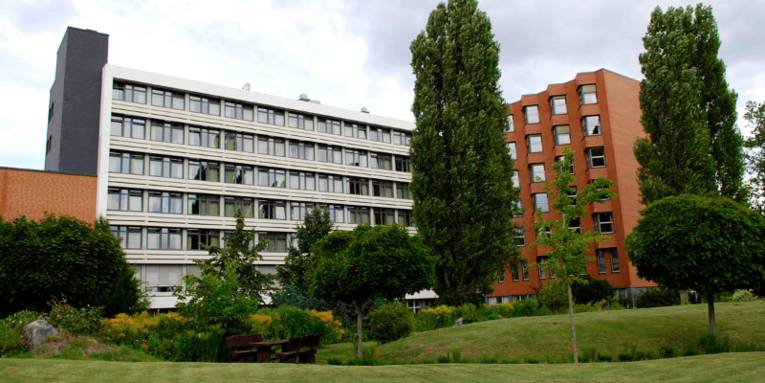 Gebäudekomplex in L-Form, der linke Teil ist weiß, der rechte, abgewinkelte Teil besteht aus roten Backsteinen. Vor dem Gebäude ist eine parkähnliche Fläche mit Rasen, Bäumen und Sträuchern zu erkennen.