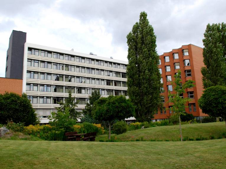 Gebäudekomplex in L-Form, der linke Teil ist weiß, der rechte, abgewinkelte Teil besteht aus roten Backsteinen. Vor dem Gebäude ist eine parkähnliche Fläche mit Rasen, Bäumen und Sträuchern zu erkennen.