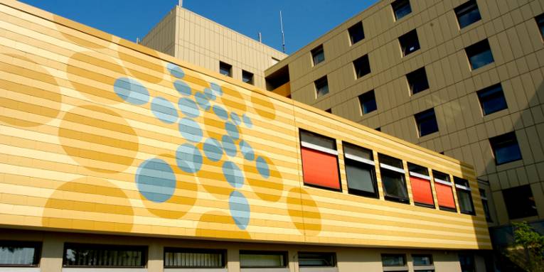 Fassade des Klinikgebäudes in Neustadt am Rübenberge. Die Aufnahme bildet eine Stelle ab, an der mehrere Gebäudeteile ineinander übergehen. Das vorderste Vauteil ist mit gelber Farbe gestrichen, darauf ist das blaue Punktelogo der Region Hannover zu erkennen.