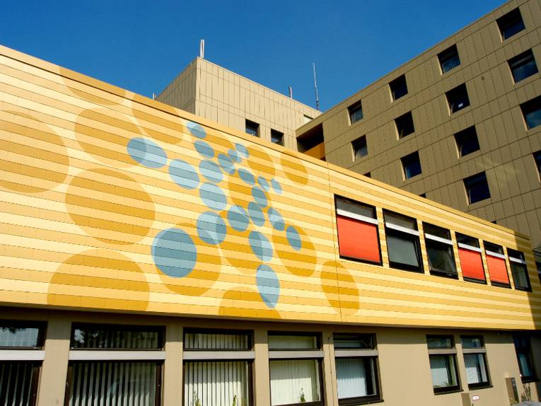 Fassade des Klinikgebäudes in Neustadt am Rübenberge. Die Aufnahme bildet eine Stelle ab, an der mehrere Gebäudeteile ineinander übergehen. Das vorderste Vauteil ist mit gelber Farbe gestrichen, darauf ist das blaue Punktelogo der Region Hannover zu erkennen.