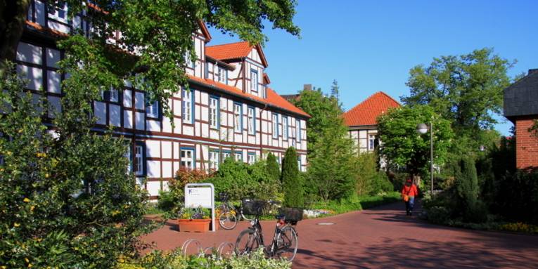 Großes Fachwerkgebäude über zweieinhalb Geschosse, entlang der gepflasterten Wege sind parkähnliche Beete mit unterschiedlich hohen Pflanzen. Ein Fahrrad ist vor dem Gebäude abgestellt, ein Fußgänger benutzt den Weg.
