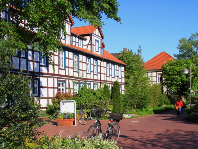 Großes Fachwerkgebäude über zweieinhalb Geschosse, entlang der gepflasterten Wege sind parkähnliche Beete mit unterschiedlich hohen Pflanzen. Ein Fahrrad ist vor dem Gebäude abgestellt, ein Fußgänger benutzt den Weg.