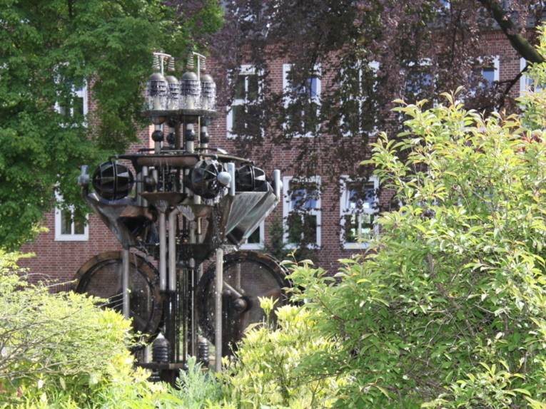 Blick durch grünes Buschwerk an einem Brunnen aus Stahl vorbei auf das Klinikgebäude. Die Fassade besteht aus roten Klinkern, die Fenster haben weiße Rahmen.